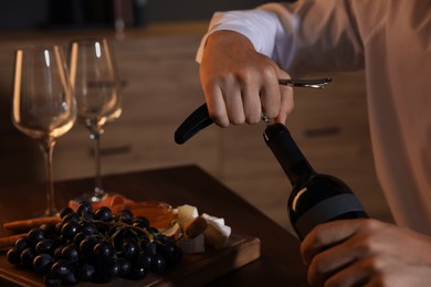 Romantic dinner. Man opening wine bottle with corkscrew at table indoors, closeup