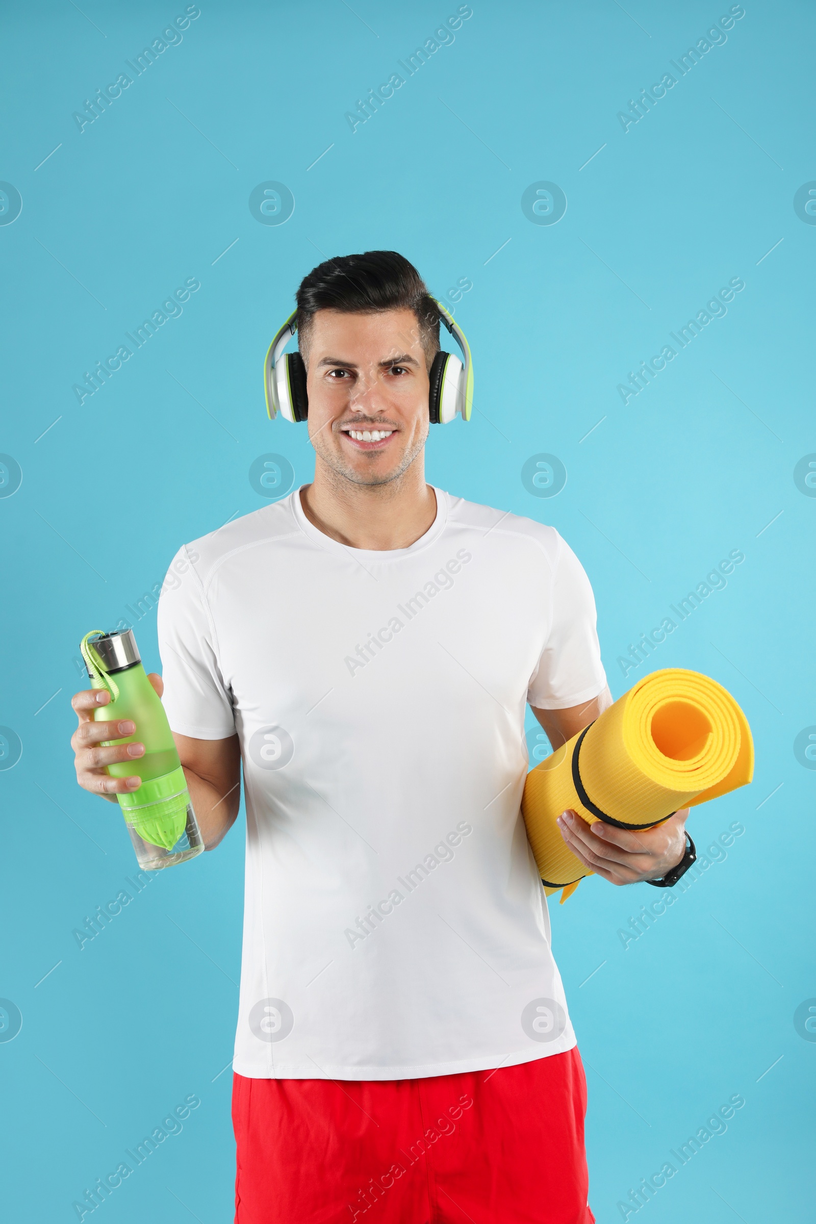 Photo of Handsome man with yoga mat, headphones and bottle on turquoise background