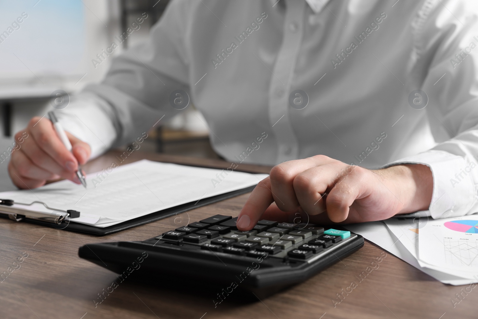 Photo of Man using calculator while taking notes at wooden table indoors, closeup