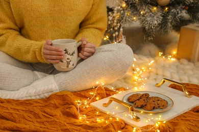 Woman with cup of hot drink and cookies sitting near Christmas tree on floor in room, closeup