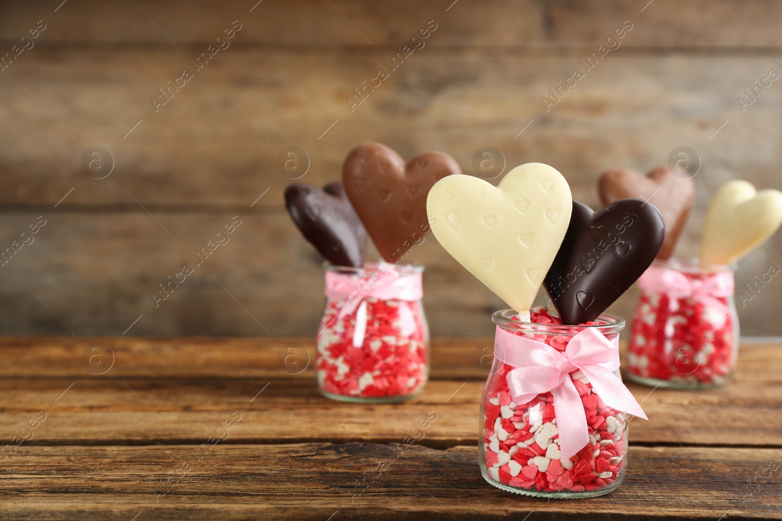 Photo of Heart shaped lollipops made of chocolate with sprinkles in glass jars on wooden table, space for text