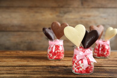 Heart shaped lollipops made of chocolate with sprinkles in glass jars on wooden table, space for text