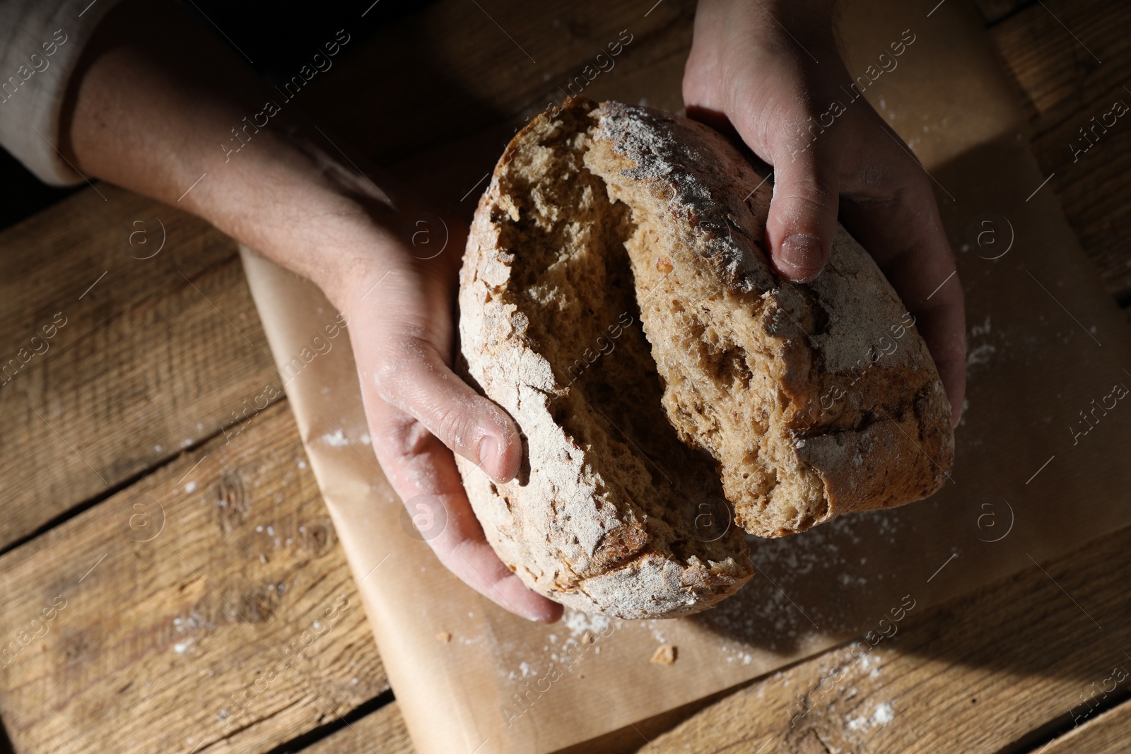 Photo of Man breaking loaf of fresh bread at wooden table, top view