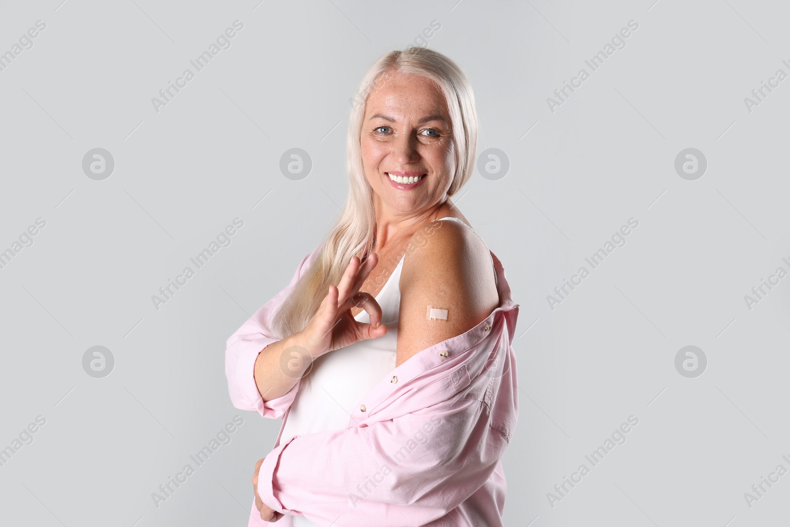 Photo of Happy mature woman showing arm with bandage after vaccination and okay gesture on light background