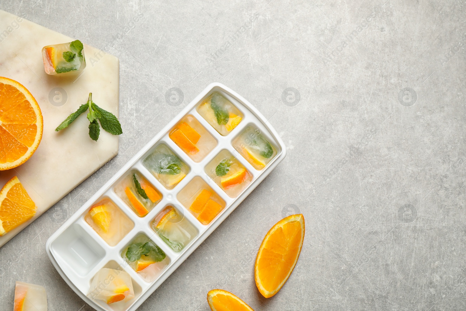 Photo of Ice cubes in tray with orange slices and mint on light grey table, flat lay