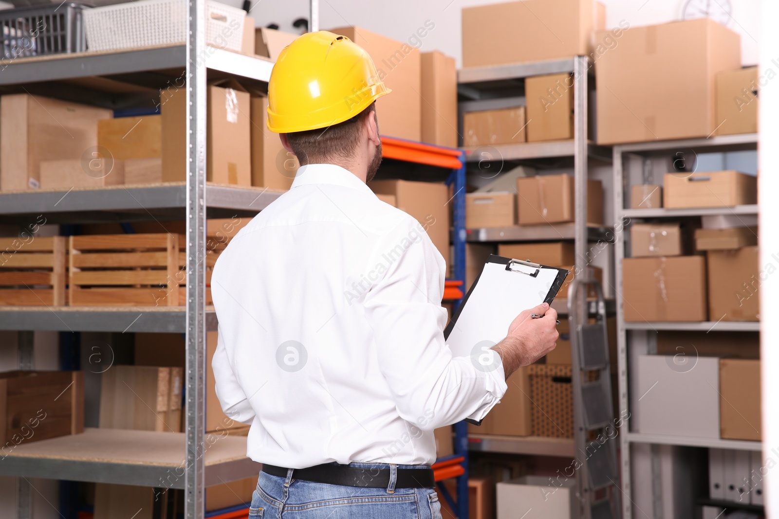 Photo of Young man with clipboard near rack of cardboard boxes at warehouse, back view