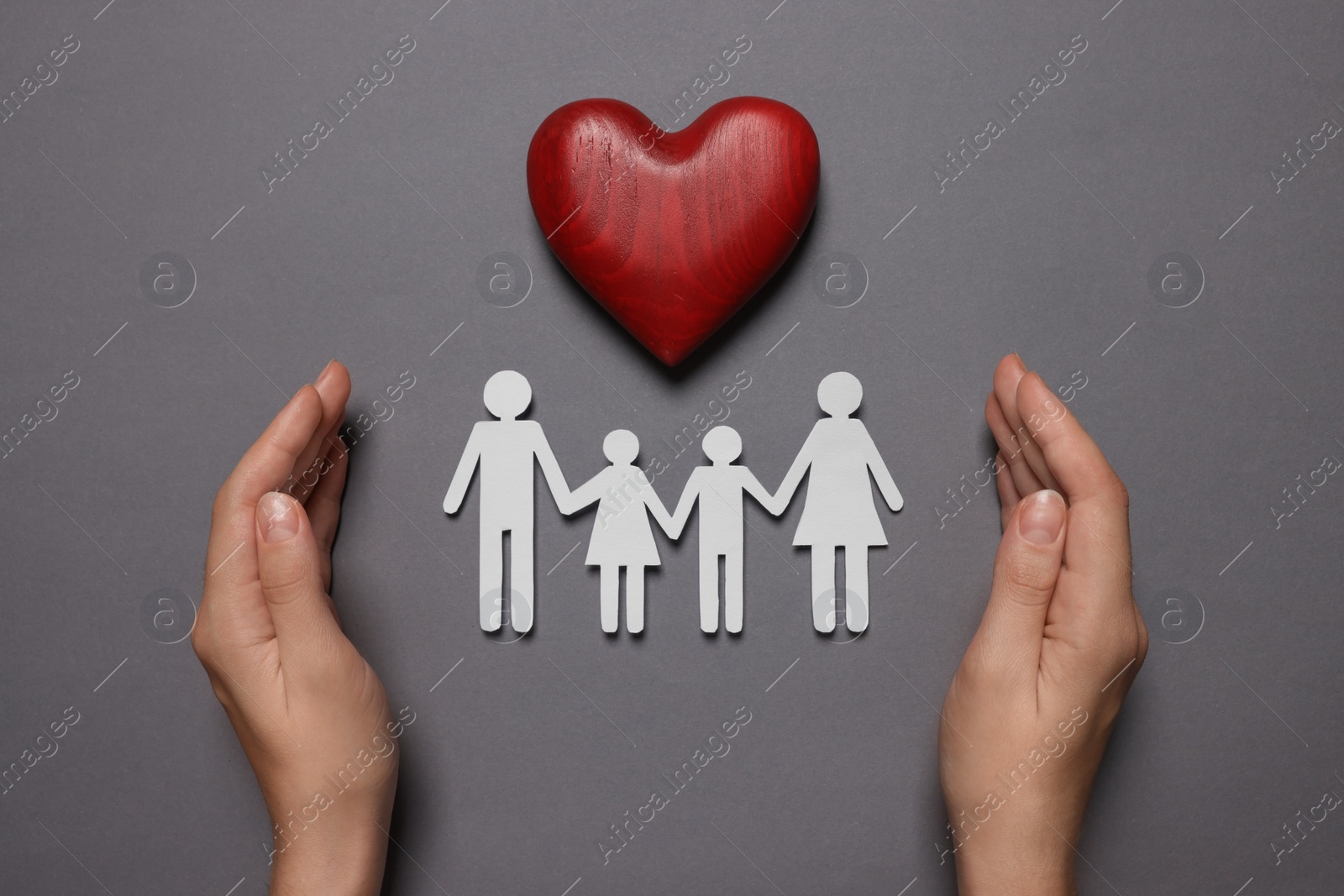 Photo of Woman protecting paper family figures and red wooden heart on grey background, top view. Insurance concept