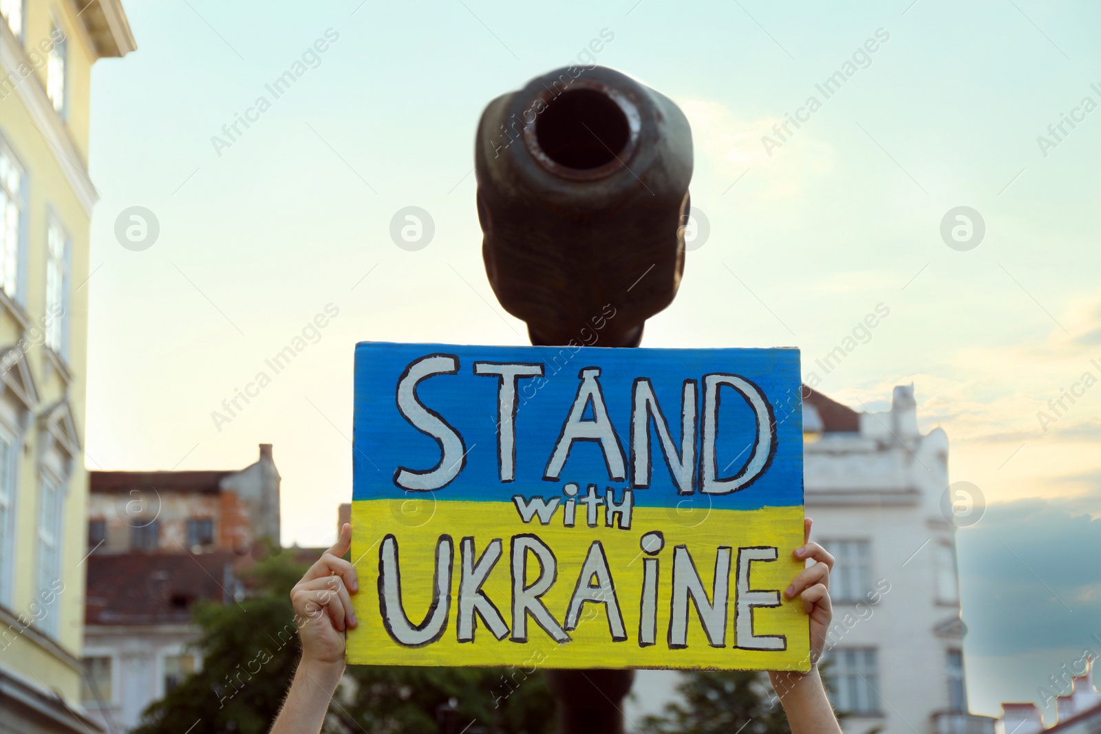 Photo of Woman holding poster in colors of national flag and words Stand With Ukraine near broken military tank outdoors, closeup