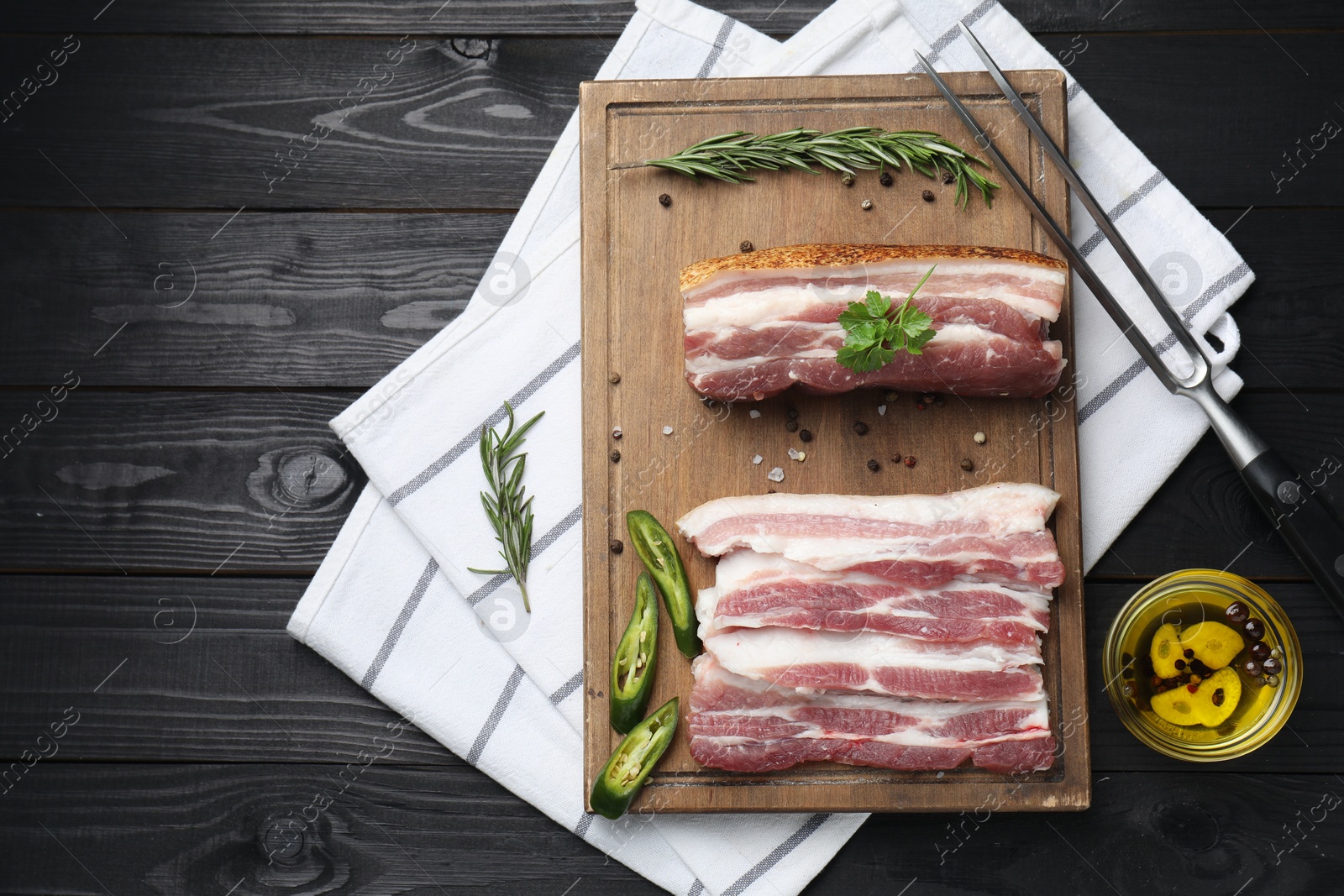 Photo of Pieces of raw pork belly, chili pepper, peppercorns, rosemary and parsley on black wooden table, flat lay