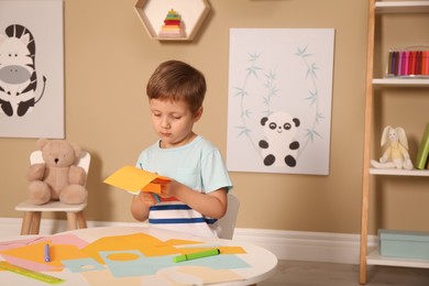 Little boy cutting color paper with scissors at table indoors