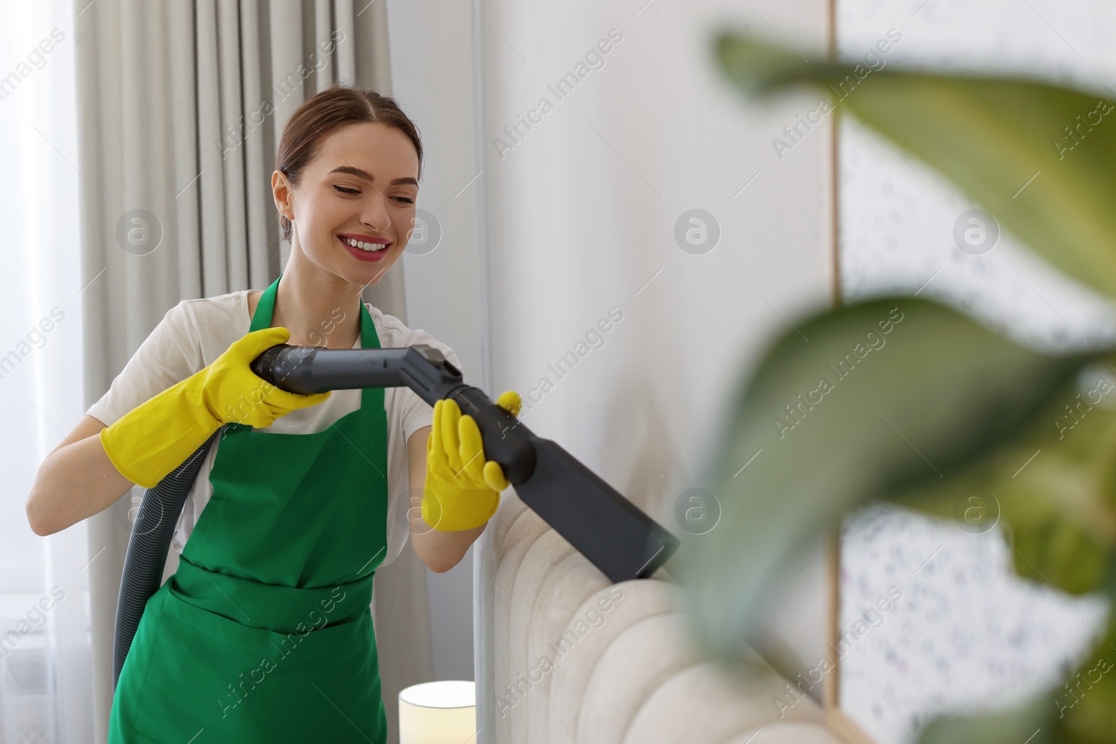 Photo of Professional janitor in uniform vacuuming bed indoors