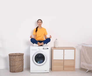 Photo of Young woman meditating on washing machine at home. Laundry day
