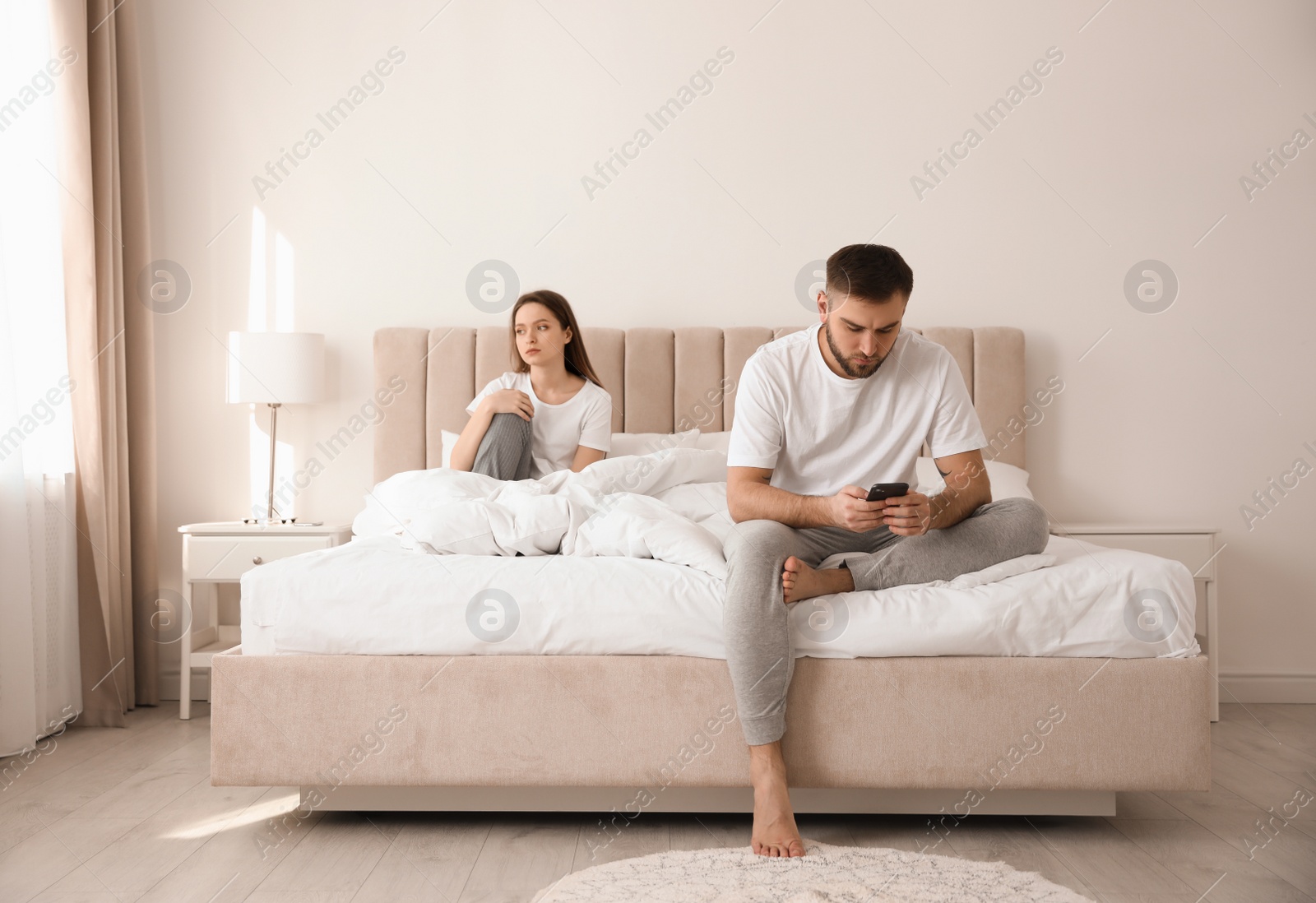 Photo of Young man preferring smartphone over his girlfriend on bed at home. Relationship problems