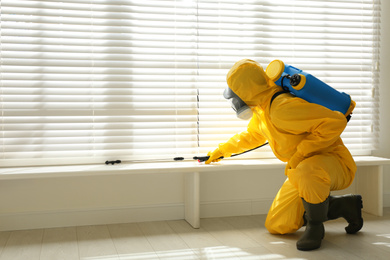 Photo of Male worker in protective suit spraying insecticide on window sill indoors, space for text. Pest control
