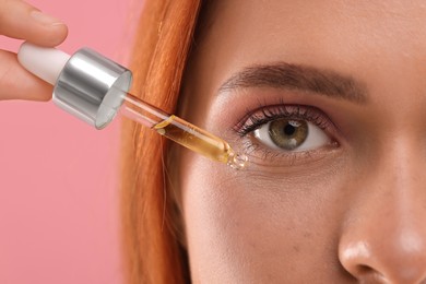 Beautiful young woman applying cosmetic serum onto her face on pink background, closeup