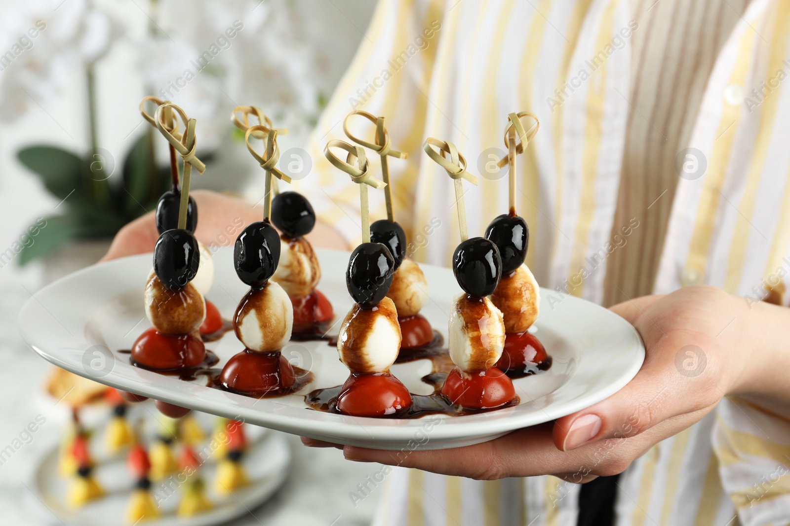 Photo of Woman holding plate of tasty canapes with black olives, mozzarella and cherry tomatoes indoors, closeup