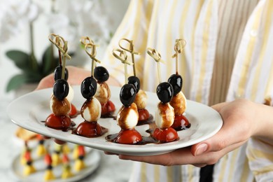 Photo of Woman holding plate of tasty canapes with black olives, mozzarella and cherry tomatoes indoors, closeup