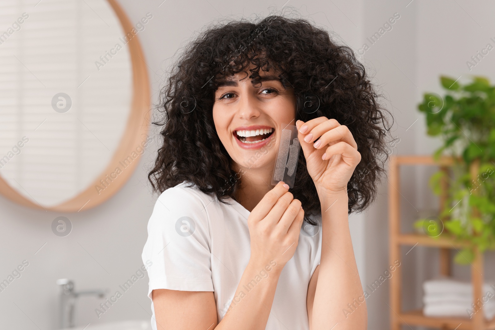 Photo of Young woman holding teeth whitening strips in bathroom