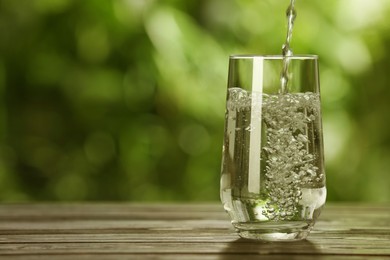 Photo of Pouring water into glass on wooden table outdoors, space for text