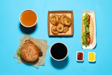 Photo of Tasty burger, hot dog, fried onion rings, different sauces and refreshing drinks on light blue background, flat lay. Fast food