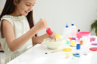 Photo of Cute little girl mixing ingredients with silicone spatula at table in room. DIY slime toy