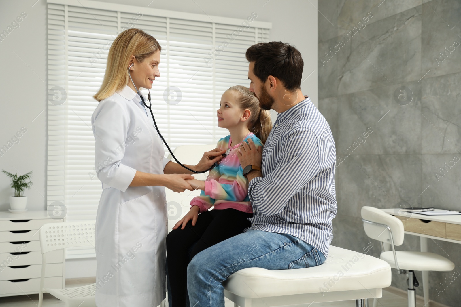 Photo of Father and daughter having appointment with doctor. Pediatrician examining little patient with stethoscope in clinic