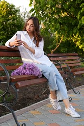 Photo of Attractive young woman with mobile phone and lilac flowers on wooden bench in park