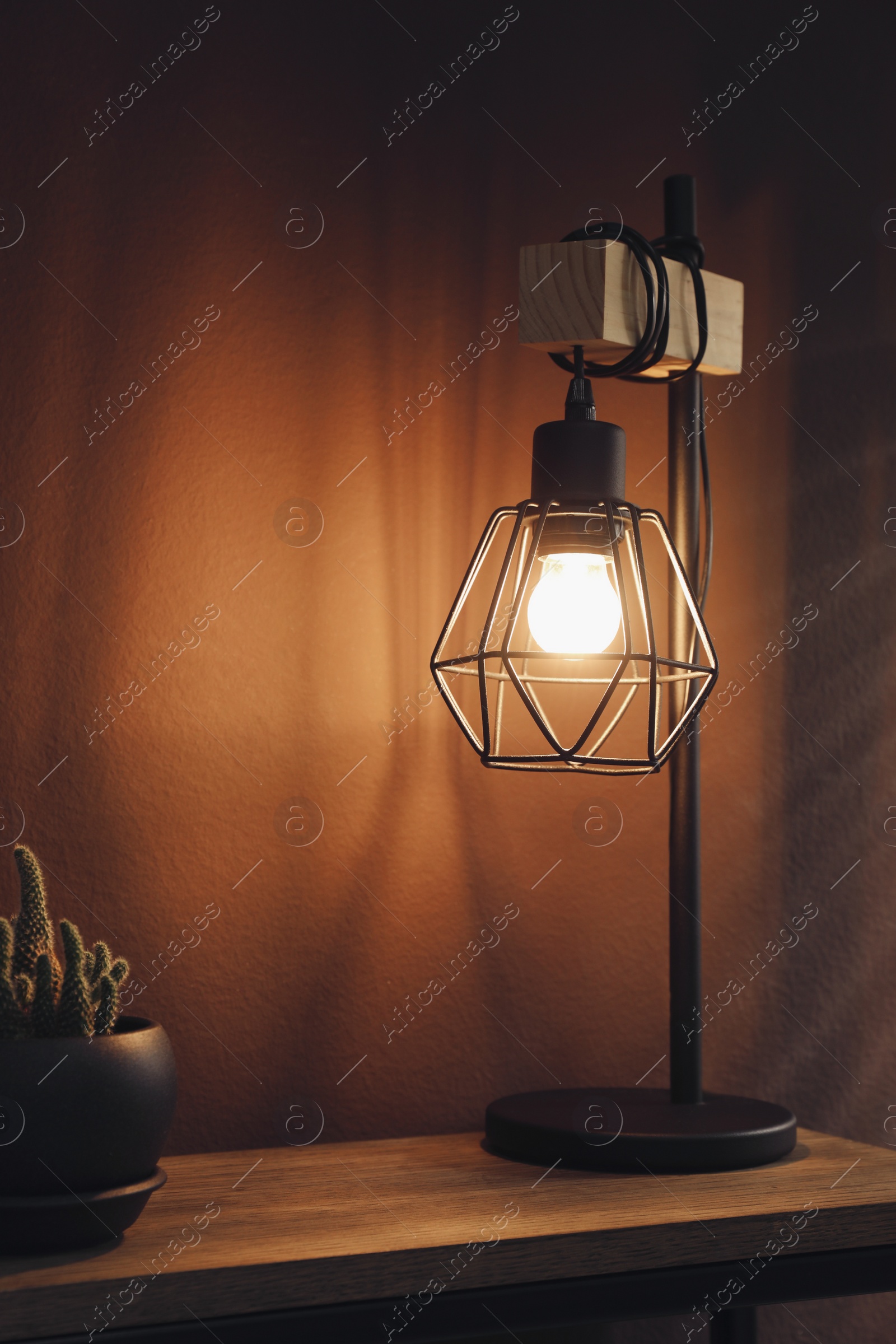 Photo of Stylish lamp, decor and green plant on wooden table near brown wall indoors. Interior design