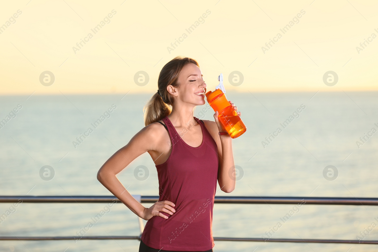 Photo of Young woman drinking water from bottle after fitness exercises on pier in morning