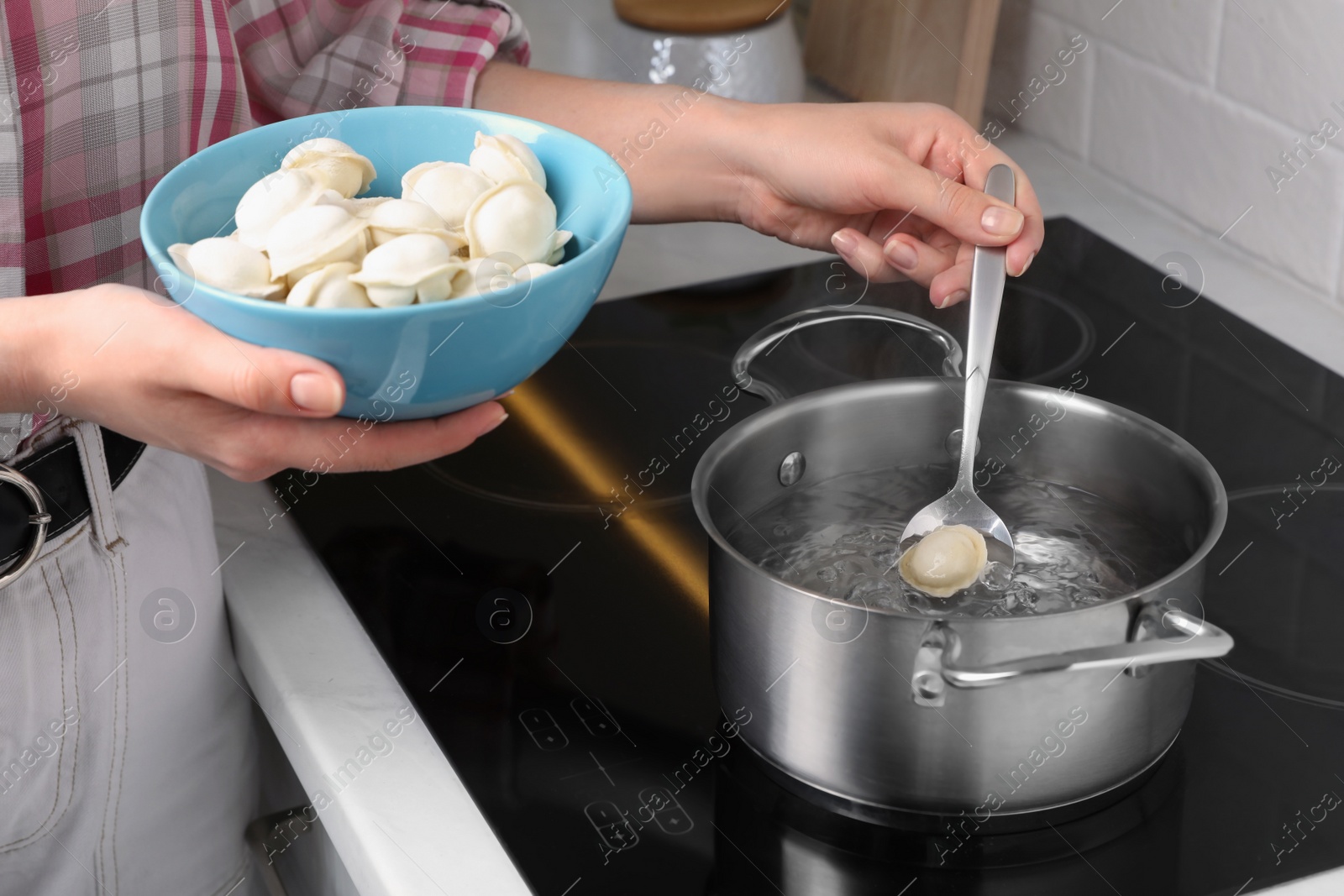 Photo of Woman putting frozen dumplings into saucepan with boiling water on cooktop in kitchen, closeup