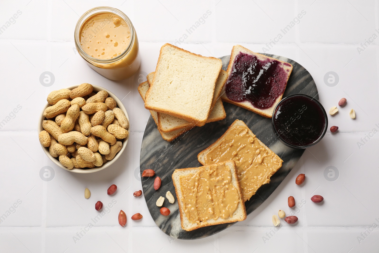 Photo of Delicious toasts with peanut butter, nuts and jam on white tiled table, flat lay