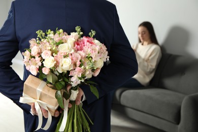 Photo of Man hiding bouquet of flowers and present for his beloved woman indoors, closeup