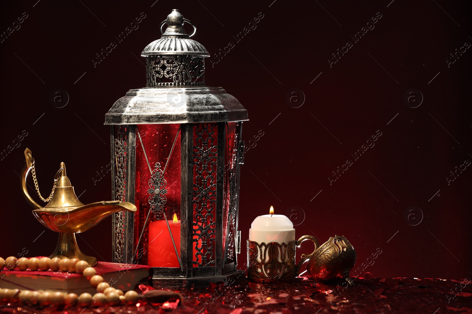 Photo of Arabic lantern, Quran, misbaha, burning candle and Aladdin magic lamp on shiny red table. Space for text