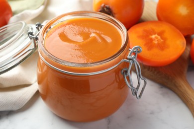 Photo of Delicious persimmon jam and fresh fruits on white marble table, closeup