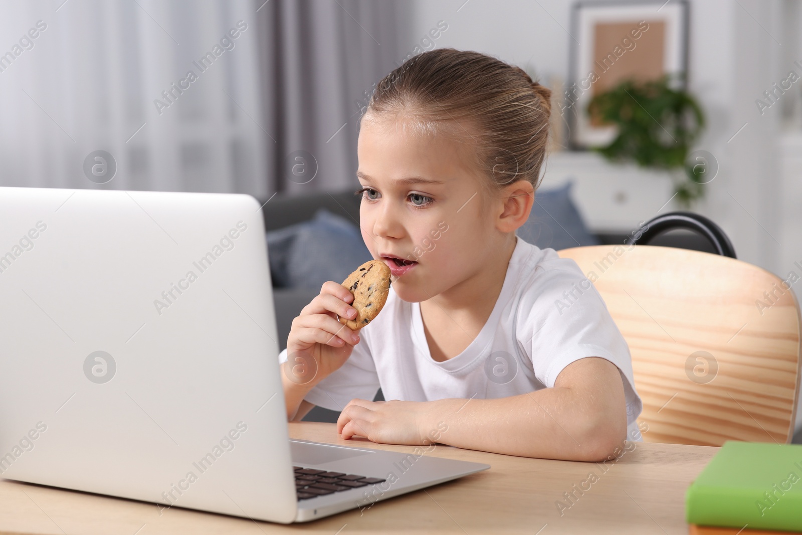Photo of Little girl using laptop and eating biscuit at table indoors. Internet addiction