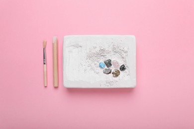 Photo of Educational toy for motor skills development. Excavation kit (plaster, wooden chisel, brush and gemstones) on pink background, flat lay