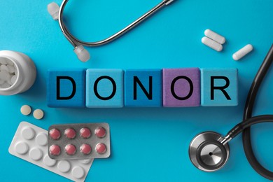 Word Donor made of wooden cubes, pills and stethoscope on light blue background, flat lay