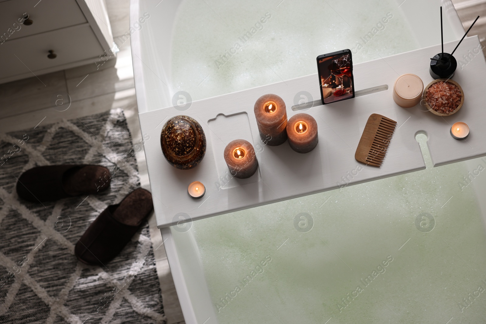 Photo of White wooden tray with smartphone, burning candles and spa products on bathtub in bathroom, above view