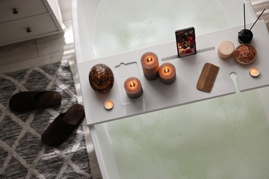 White wooden tray with smartphone, burning candles and spa products on bathtub in bathroom, above view