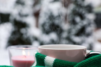 Photo of Winter morning. Cup with hot drink wrapped in green sweater and burning candle outdoors, closeup. Space for text