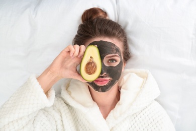Photo of Young woman with facial mask and avocado lying on bed, top view