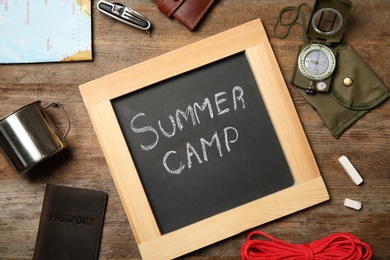 Chalkboard with text SUMMER CAMP and camping equipment on wooden background, flat lay