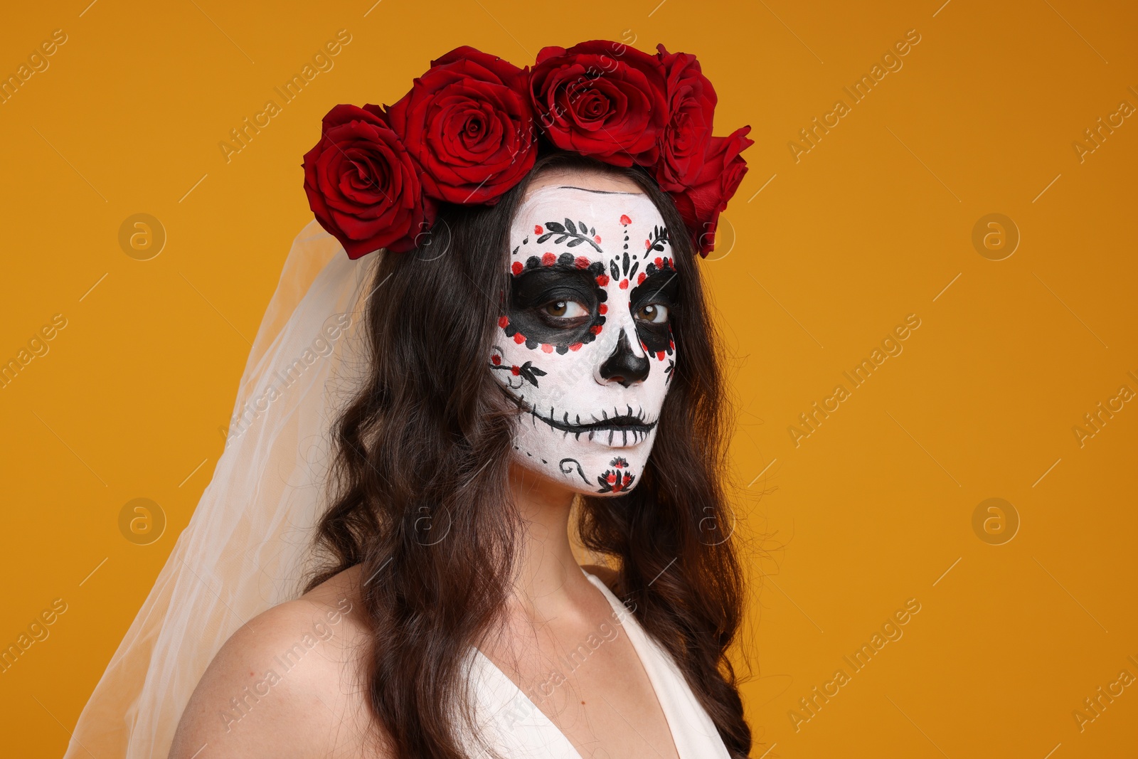 Photo of Young woman in scary bride costume with sugar skull makeup and flower crown on orange background. Halloween celebration