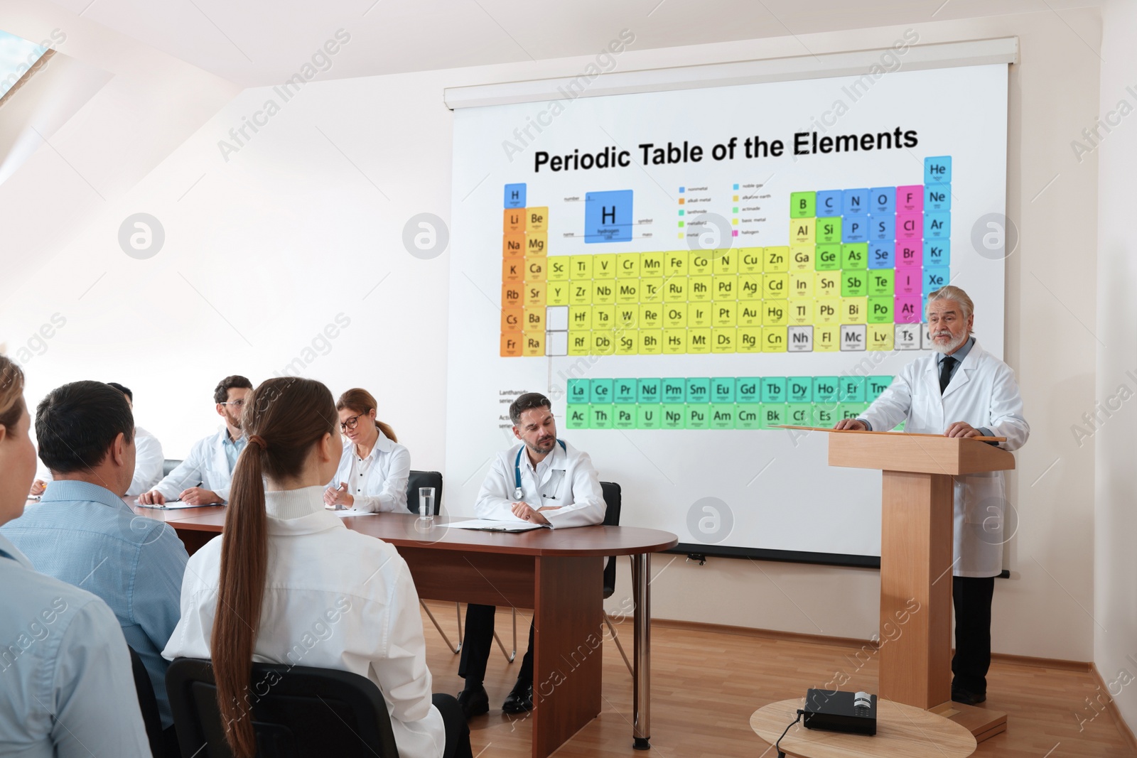 Image of Lecture in chemistry. Senior speaker near projection screen with illustration of periodic table. Professors and audience in conference room