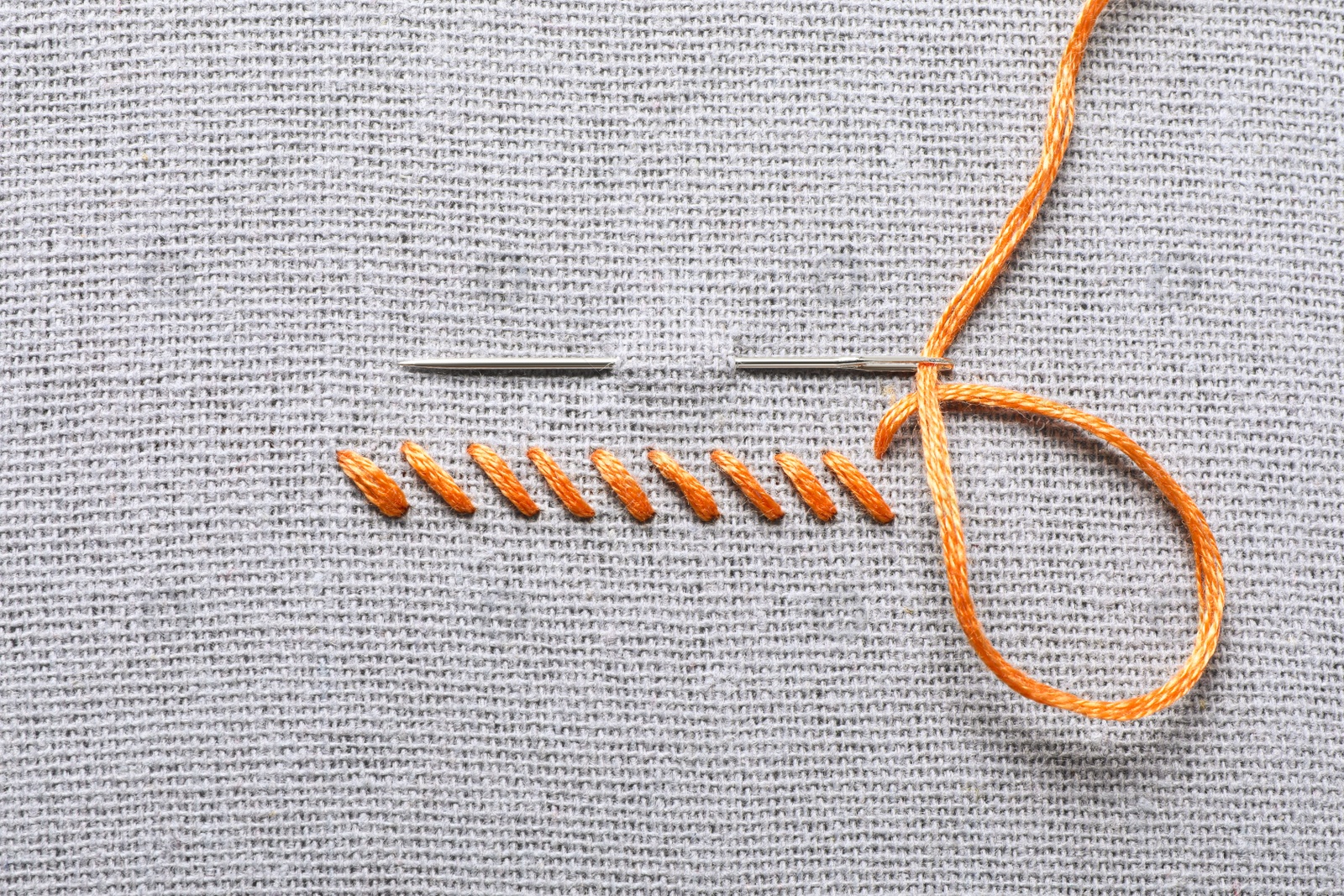 Photo of Needle with orange embroidery floss and row of stitches on grey fabric, top view