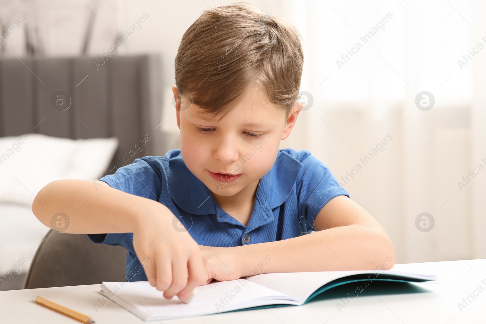 Photo of Little boy erasing mistake in his notebook at white desk indoors