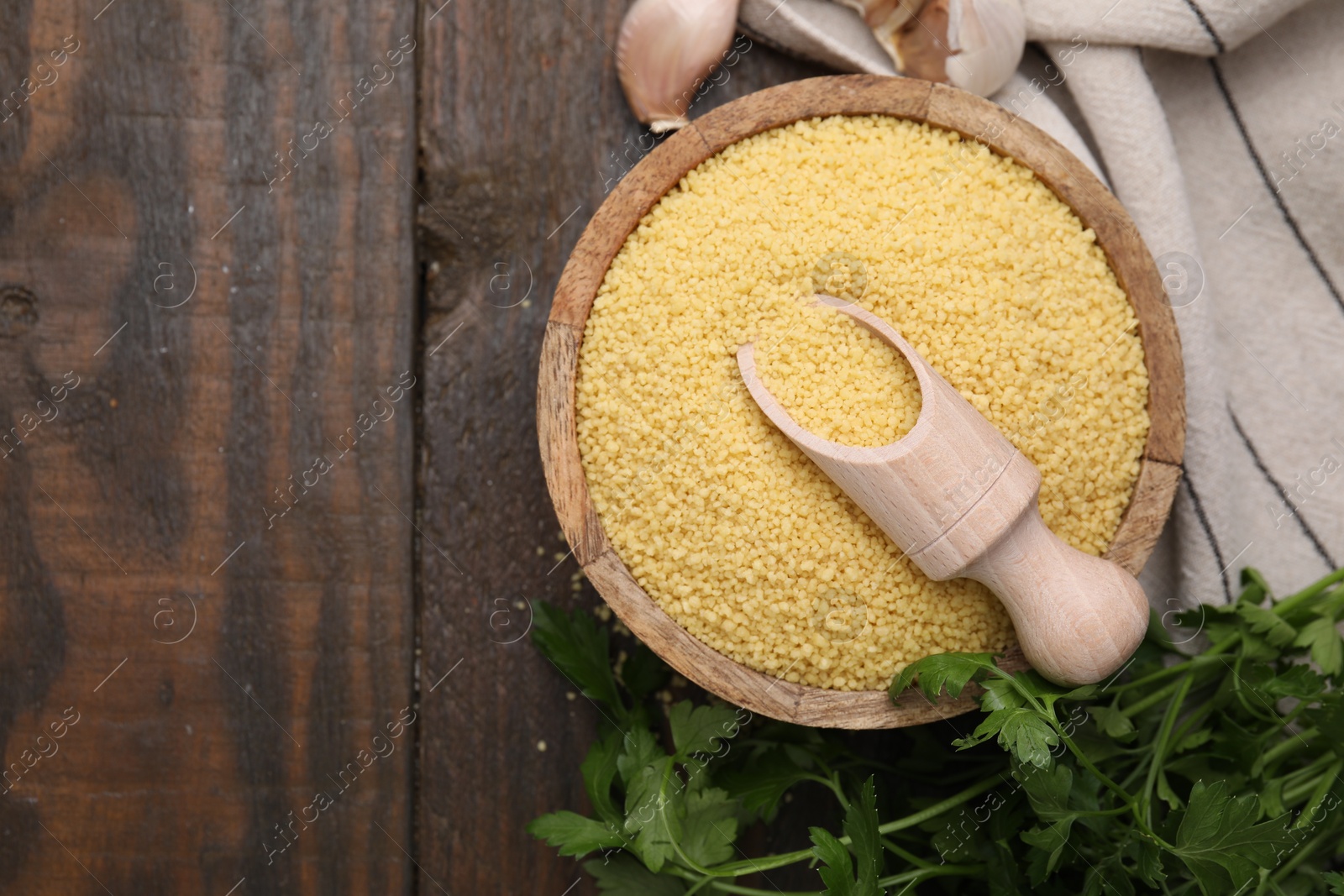 Photo of Raw couscous in bowl, scoop, parsley and garlic on wooden table, flat lay. Space for text