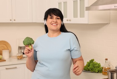 Photo of Happy overweight woman with headphones and broccoli in kitchen. Healthy diet