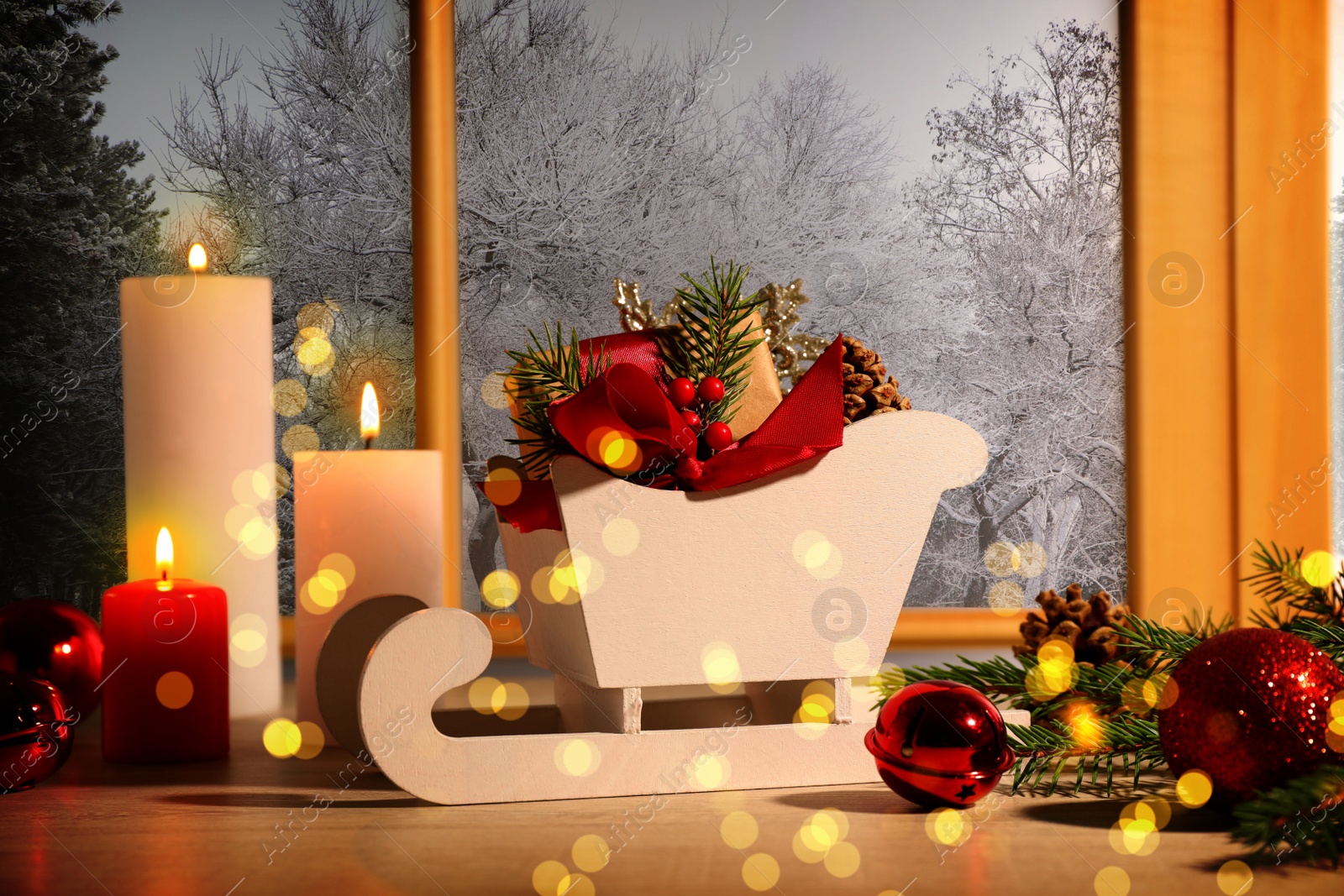 Image of Small wooden sleigh with gift box near burning candles and festive decor on window sill indoors. Christmas eve