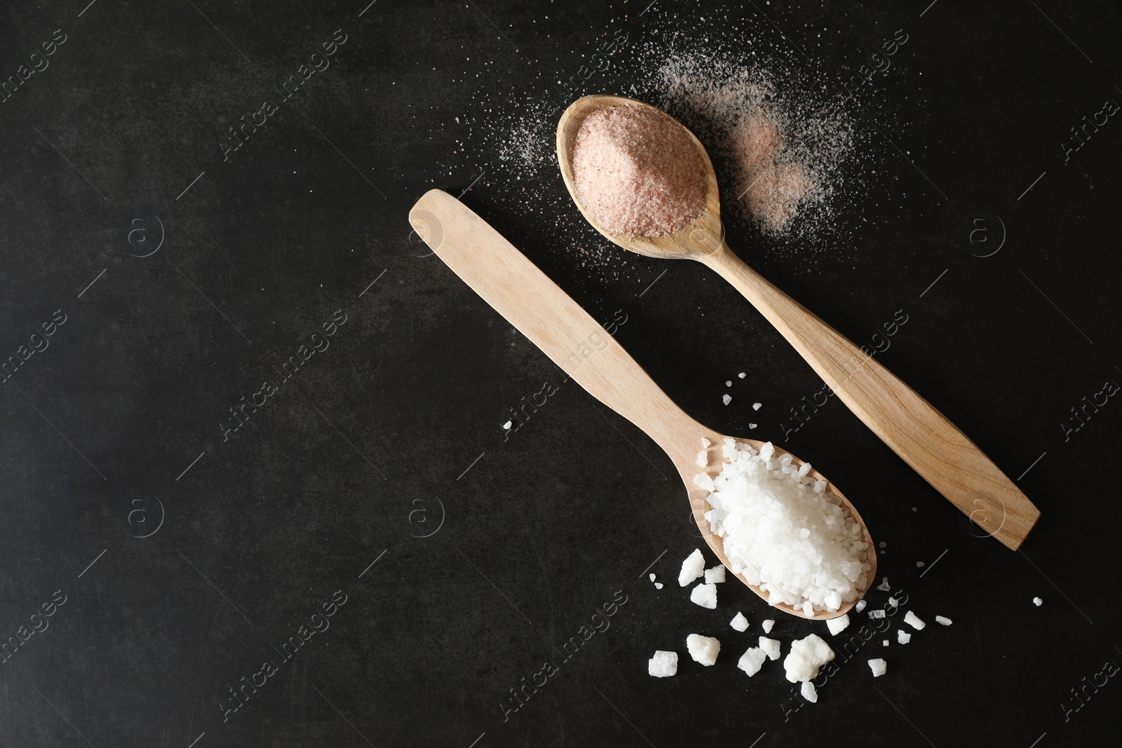 Photo of Different types of organic salt in spoons on black table, flat lay. Space for text